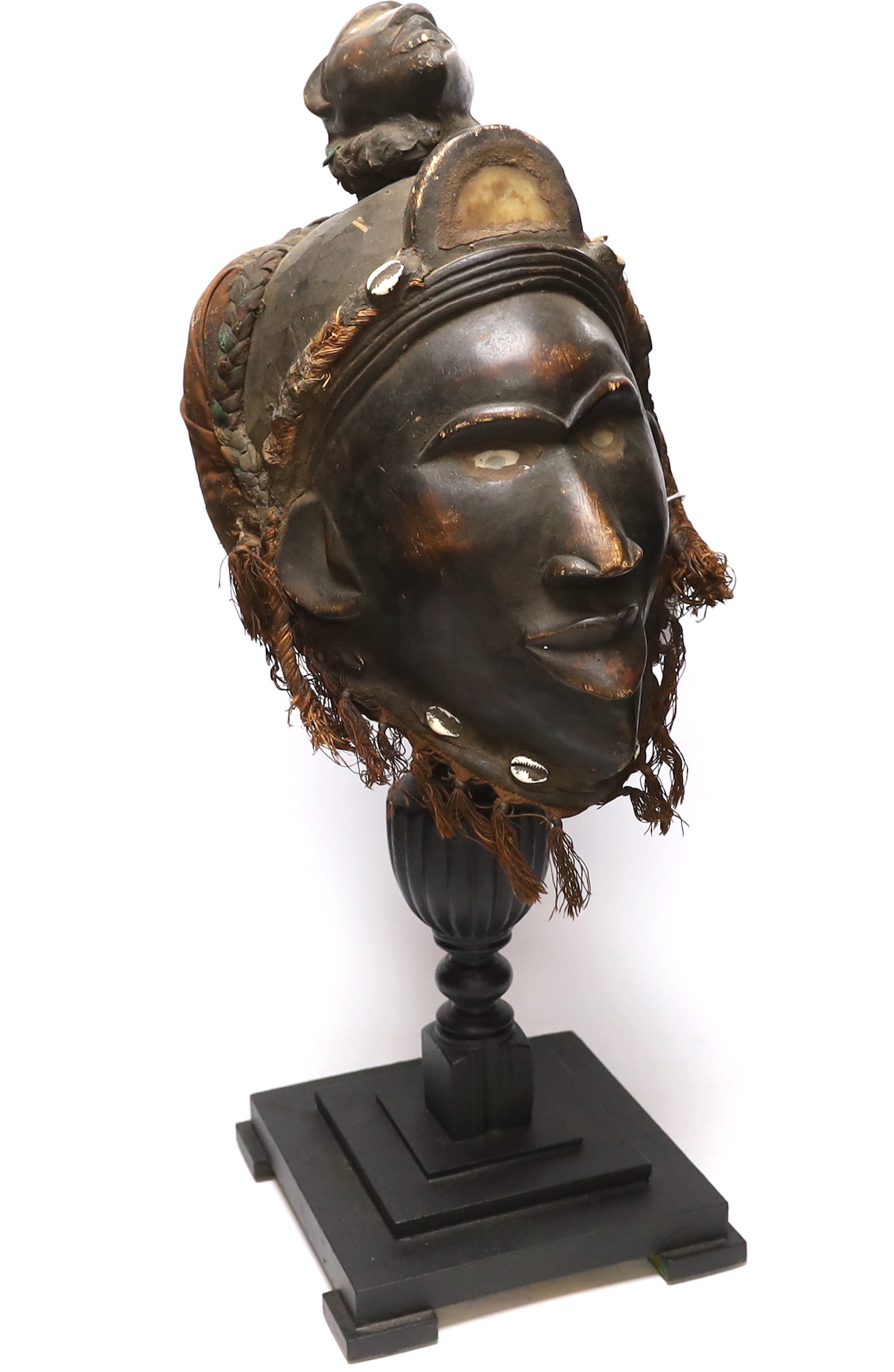 A West African carved wood helmet mask, possibly Kuba (Democratic Republic of Congo), with applied shells and glass inserts, on hardwood stand